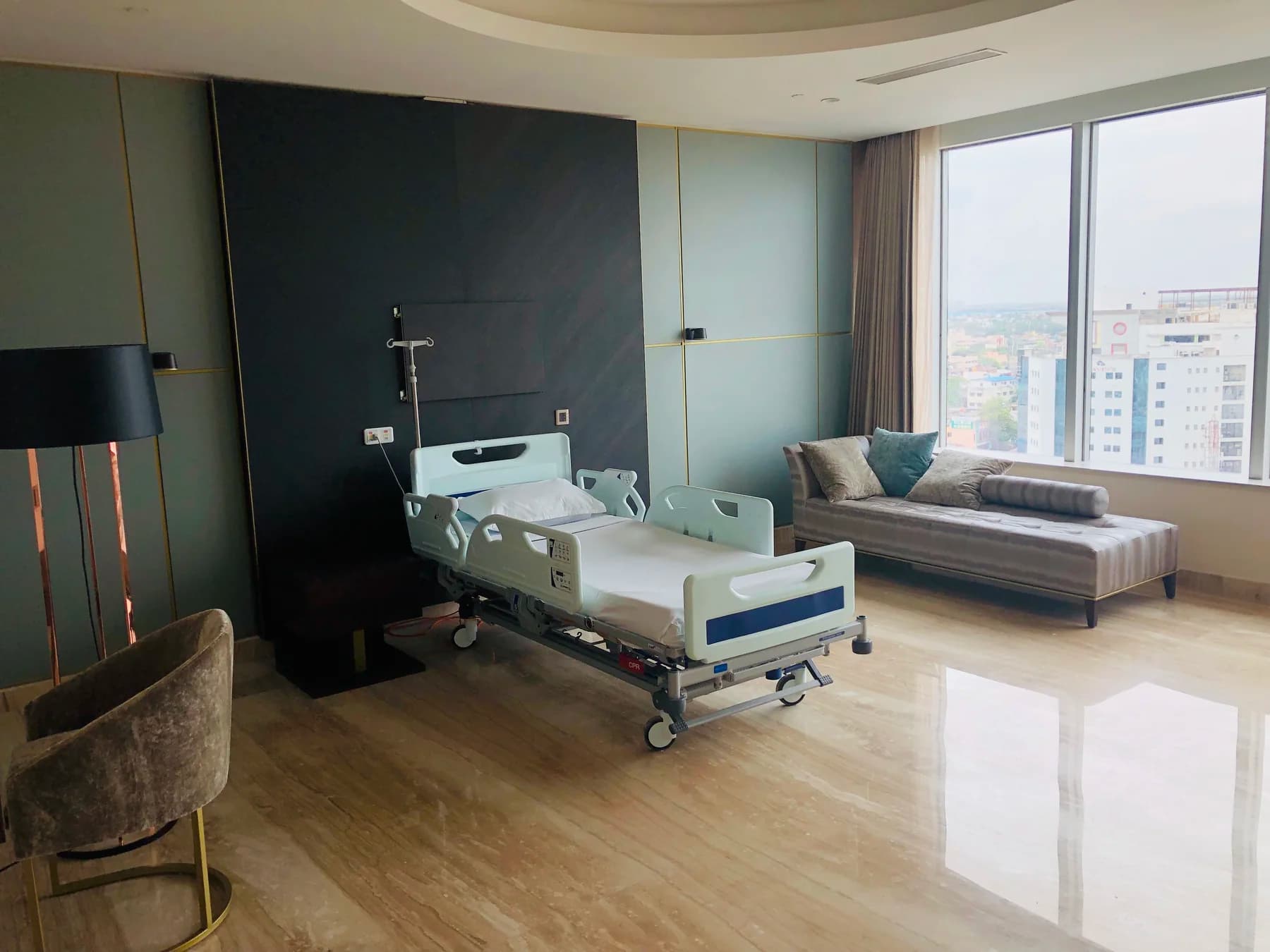 A private hospital suite with a hospital bed, day bed and floor-to-ceiling window over the city